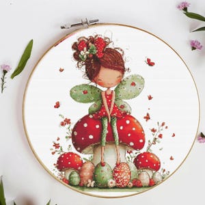 May include: Cross-stitch embroidery of a fairy girl with brown hair, red dress, and green wings, sitting on a mushroom. The fairy is surrounded by red butterflies and mushrooms with white spots, all set within a wooden embroidery hoop.