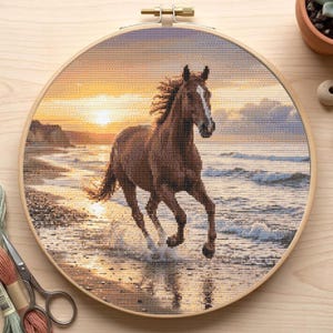 Puede incluir: Un bordado de punto de cruz de un caballo marrón corriendo en una playa al atardecer. El caballo tiene una mancha blanca y chapotea en el agua. El fondo presenta un cielo dorado y olas, enmarcado en un aro de madera.