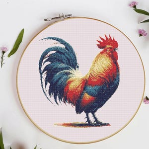 May include: A colorful cross-stitch of a rooster, featuring red, blue, and yellow hues. The rooster is displayed within a wooden embroidery hoop against a light pink background. The artwork is detailed and vibrant, showcasing the bird's feathers and form.
