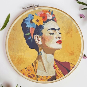 May include: Cross-stitch portrait of a woman with closed eyes, wearing a floral crown and earrings, set in a wooden embroidery hoop. The background is a textured yellow, and the woman has red lipstick and a patterned top.