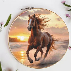 Puede incluir: Un bordado de punto de cruz de un caballo marrón galopando en una playa al atardecer. El caballo tiene una mancha blanca y una melena fluida. El fondo presenta un cielo y un océano de color naranja y amarillo vibrantes. El bordado está en un aro de madera redondo.