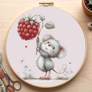 May include: A cross-stitch embroidery hoop featuring a cute cartoon mouse holding a large red raspberry. The mouse is gray with pink accents, set against a light pink background with floral details. The wooden hoop is visible.