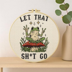 May include: A circular wooden embroidery hoop containing a cross-stitch design of a green frog sitting on a red mushroom with white spots. The frog is making peace signs with both hands. The text LET THAT SH*T GO is stitched in black.