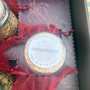 Könnte beinhalten: Ein Glasgefäß mit getrockneten Teeblättern, einem Korkverschluss und einem runden Etikett mit der Aufschrift SUTCHATEAS CRONES SHADOW THE RED THREAD. Das Glas befindet sich in einer dunklen Schachtel mit einem dunkelroten Organzaband und Einlage.
