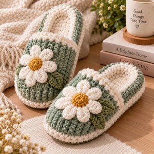 May include: A pair of sage green and cream crochet slippers featuring a large white daisy flower with a yellow center on the top of each slipper. The slippers have a thick, textured knit construction with a soft cream-colored interior lining.