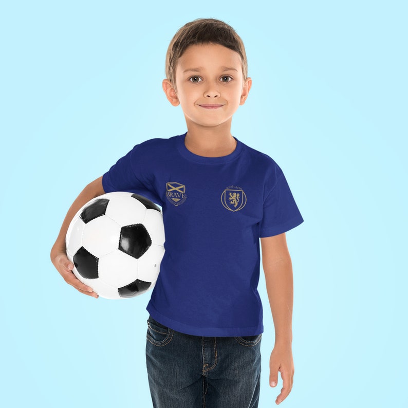 May include: A young boy wearing a blue t-shirt with a gold embroidered logo that says "Brave" and a Scottish crest. He is holding a white and black soccer ball.