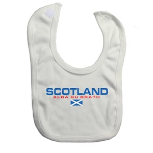 Puede incluir: Babero blanco para bebé con la palabra "SCOTLAND" en letras azules en bloque sobre "ALBA GU BRATH" en rojo. Una bandera escocesa azul y blanca está debajo. El babero tiene un cierre de velcro.
