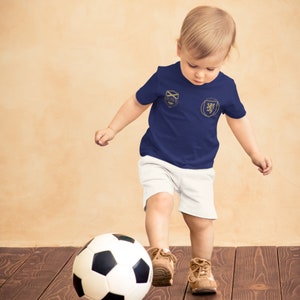 May include: A young child wearing a navy blue t-shirt with gold embroidery of a crest and a lion. The child is kicking a black and white soccer ball with their right foot. The child is wearing white shorts and brown shoes.
