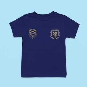 May include: A navy blue t-shirt with a gold Scottish crest on the right side and a gold crest with the word "BRAVE" on the left side.