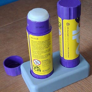 May include: Two purple glue sticks with yellow labels, one open with a white adhesive tip and one closed with a cap, standing in a light grey rectangular holder. The labels feature text including 40g e, ypo.co.uk, and a warning symbol for children under 36 months.