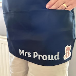 Könnte beinhalten: Eine schwarze Taillenschürze mit einer großen Vordertasche und dem silbernen Glitzertext Mrs Proud auf der Unterseite. Eine kleine Grafik eines braunen Teddybären befindet sich rechts neben dem Text in der unteren Ecke der Schürze.