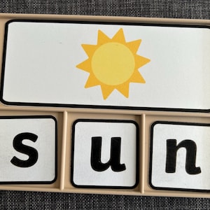 May include: A beige plastic tray containing four white rectangular tiles. The top tile features a yellow sun illustration. The three bottom tiles each display a single black lowercase letter, spelling out the word sun in sequence.