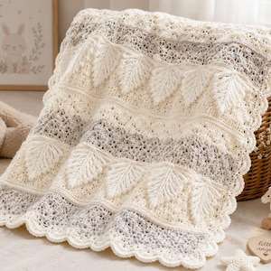 Könnte beinhalten: Eine cremefarbene und hellgraue gehäkelte Decke mit einem wiederkehrenden Muster aus erhabenen Blattmotiven, komplizierten Lochstichmustern und einem gewellten Rand. Das Textil zeigt abwechselnde horizontale Bänder aus festen und spitzenartigen Strukturen.