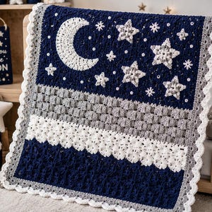 Op de afbeelding: Een rechthoekige gehaakte deken met een donkerblauw bovengedeelte met een grote witte halve maan en diverse sterren. Het middengedeelte heeft een gestructureerd grijs mandvlechtpatroon en een witte bloemenrand met een geschulpte afwerking.