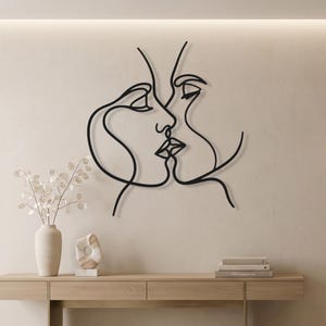 Puede incluir: Arte mural de metal negro que representa dos rostros en una línea continua, besándose. La obra de arte está montada en una pared beige sobre una consola de madera clara. Un jarrón blanco con ramas decorativas y una pequeña escultura están sobre la mesa.