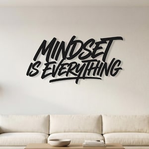 Könnte beinhalten: Ein schwarzes Wandobjekt mit dem Schriftzug "MINDSET IS EVERYTHING" in einer fetten, stilisierten Handschrift. Die Buchstaben sind miteinander verbunden und haben ein dickes, dunkles Erscheinungsbild, das ein markantes visuelles Statement setzt.