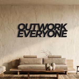以下が含まれることがあります： 太字のサンセリフ体で「OUTWORK EVERYONE」という文字が記された黒い壁掛けサイン。文字は2段に配置されており、背後にある質感のある明るい色の壁面に対して鮮明なコントラストを生み出しています。