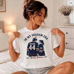 Könnte beinhalten: Weißes T-Shirt mit dem Text "IN MY GRAHAM ERA" und einer Grafik eines Paares in Hockey-Trikots. Die marineblauen Trikots zeigen den Namen "GRAHAM" und die Nummer "44". Der Schriftzug "Hockey Romance" befindet sich darunter.