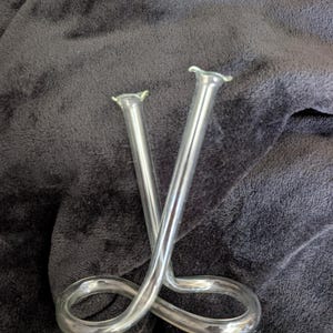 May include: Clear glass bud vase with a unique looped design. The vase has two slender, upright tubes that cross over each other, connected by a curved base. The vase is made of clear glass and is suitable for displaying small flowers or stems.