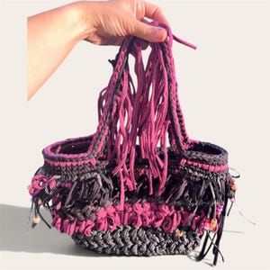 Può includere: Una borsa all'uncinetto fatta a mano in filato di tessuto grigio scuro e magenta. La borsa presenta una base intrecciata testurizzata, un corpo decorato con lunghe strisce di frange e due manici integrati. Piccole perline di plastica colorate accentuano le frange.