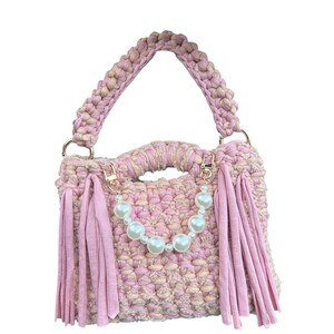 Könnte beinhalten: Handgefertigte, gehäkelte Handtasche in Rosa und Beige mit geflochtenem Henkel und goldfarbenen Beschlägen. Die Tasche hat rosa Fransen und eine Perlenkette. Ein stilvolles Accessoire.