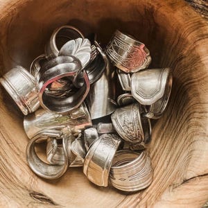 May include: A wooden bowl filled with silver spoon rings. The rings, crafted from repurposed silverware, display a variety of designs and patterns. The collection includes rings with ornate detailing and others with a more streamlined appearance, creating a unique assortment.