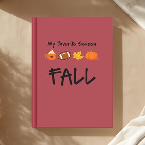 Può includere: Un taccuino con copertina rigida color bordeaux con il testo nero My Favorite Season FALL. La copertina presenta piccole icone grafiche di una tazza di caffè con un cuore, un pallone da football, una foglia d'acero gialla e una zucca arancione.