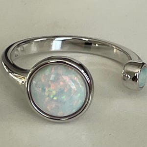 May include: A silver-toned open ring featuring two round, bezel-set opals. The larger opal is positioned at the front, while a smaller opal sits at the end of the band. The metal band has a polished finish and displays the hallmark 925 inside.