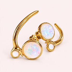 May include: A pair of gold-toned ear climbers featuring a curved metal frame set with two round, iridescent white opals of varying sizes on each piece. The smooth, polished gold metal creates a sweeping arc that follows the ear shape.