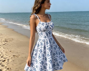 May include: A white summer dress adorned with a blue floral print. The dress has delicate spaghetti straps and a fitted bodice that transitions into a flared skirt. The dress is worn on a sandy beach with the sea visible.