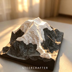 May include: A three-dimensional topographic model of a mountain with a white peak and dark gray base. The sculpture features textured slopes and ridges, with the text STEERCRAFTER printed in white at the bottom center of the rectangular base.
