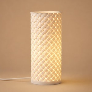 May include: A cylindrical table lamp featuring a white exterior with a repeating woven lattice pattern. The lamp emits a warm, soft glow from within, highlighting the intricate crisscross design of the structure and a white power cord.