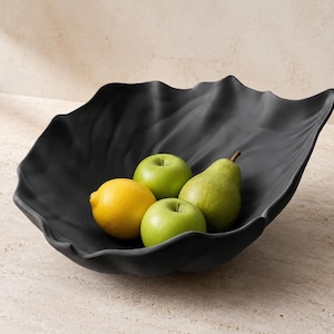 Peut inclure: Un bol noir mat avec une forme organique ondulée rappelant une feuille, contenant deux pommes vertes, un citron jaune et une poire verte. Le bol présente un bassin profond et incurvé avec des bords irréguliers qui imitent le feuillage.