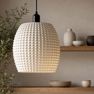 Könnte beinhalten: Cremefarbene Pendelleuchte mit strukturiertem, geometrischem Muster. Die Leuchte hängt an einem schwarzen Kabel und einer Halterung. Die Lampe ist kugelförmig und hat eine Reihe kleiner, erhabener, quadratischer Designs auf ihrer Oberfläche. Der Hintergrund umfasst Holzregale und dekorative Keramik.