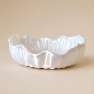 Könnte beinhalten: Eine weiße Schale mit perlmuttartigem Glanz und einem gewellten, muschelförmigen Rand. Die Innen- und Außenflächen weisen vertikale Rillen auf, die ein gefaltetes, blütenartiges Aussehen erzeugen und dem Objekt eine glatte, reflektierende Textur verleihen.