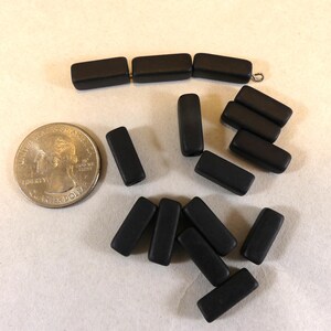 May include: A collection of matte black rectangular beads, some strung together, alongside a U.S. quarter for size comparison. The beads are smooth and evenly colored, suitable for jewelry making.
