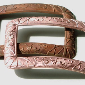 May include: Two antique metal belt buckles with ornate designs. The top buckle is a darker copper color, while the bottom buckle is a lighter, rose gold color.