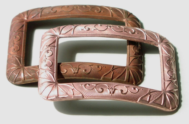 May include: Two antique metal belt buckles with ornate designs. The top buckle is a darker copper color, while the bottom buckle is a lighter, rose gold color.