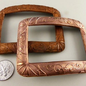 May include: Two antique copper-colored metal belt buckles with ornate floral designs. The buckles are rectangular and have a slightly raised edge.