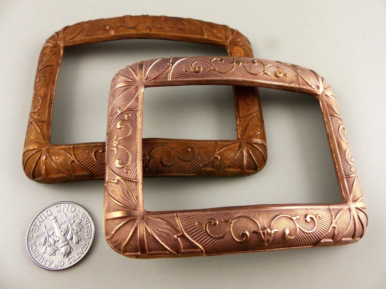 May include: Two antique copper-colored metal belt buckles with ornate floral designs. The buckles are rectangular and have a slightly raised edge.