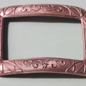 May include: A rectangular, rose gold-colored metal buckle with an ornate, engraved design.