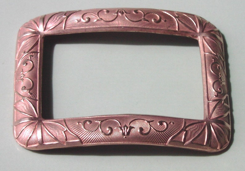 May include: A rectangular, rose gold-colored metal buckle with an ornate, engraved design.