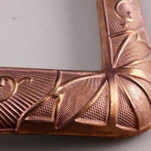 May include: Close-up of a copper-colored metal buckle with an intricate design. The buckle has a geometric pattern with a starburst design in the center.