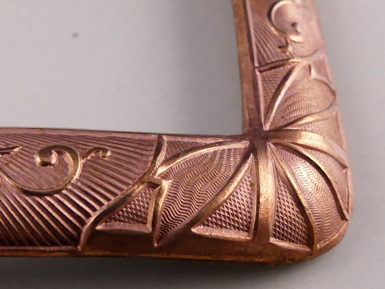 May include: Close-up of a copper-colored metal buckle with an intricate design. The buckle has a geometric pattern with a starburst design in the center.