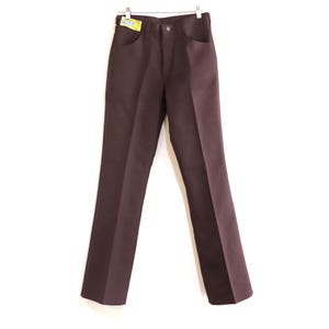 Può includere: Pantaloni marrone scuro con design a gamba dritta, appesi su sfondo bianco. I pantaloni hanno tasche anteriori e chiusura con bottoni. Una piccola etichetta è attaccata alla cintura. I pantaloni sono realizzati in tessuto liscio e tinta unita.
