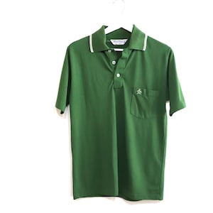 May include: Green polo shirt with a white collar trim and a chest pocket. The shirt has short sleeves and a three-button placket. A small logo is visible on the pocket. The shirt is hanging on a white hanger.