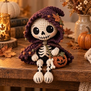 Puede incluir: Una muñeca de esqueleto tejida a ganchillo con una capa con capucha de color morado oscuro adornada con pequeños detalles naranjas y verdes y una hoja de arce. El esqueleto tiene un cuerpo blanco con cuencas negras y sostiene una calabaza.