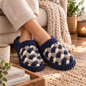 Può includere: Un paio di pantofole slip-on con un design intrecciato a maglia grossa. Le pantofole sono realizzate con filato spesso in una palette di colori blu navy, grigio chiaro e crema, che mostra un motivo intrecciato testurizzato sulla parte superiore.