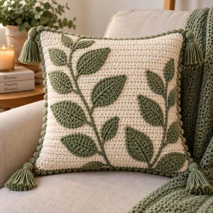 Può includere: Un cuscino decorativo quadrato all'uncinetto color crema con due motivi a foglia verde scuro cuciti sul davanti. Il cuscino presenta un bordo all'uncinetto verde scuro e una nappa abbinata attaccata a ciascuno dei quattro angoli, creando un aspetto strutturato.