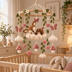 Può includere: Una giostrina per neonati all'uncinetto con un cerbiatto marrone che dorme su una nuvola bianca al centro. Include elementi pendenti come farfalle rosa, viti verdi, nuvole bianche, cuori rosa e funghi rossi con puntini bianchi su un anello di legno.